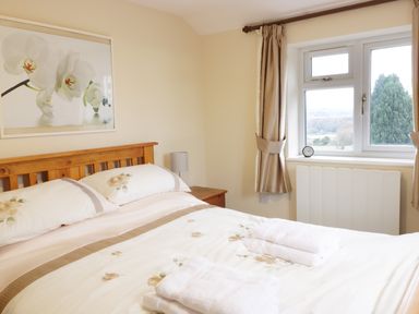 A bedroom with a wooden bed, floral bedding, folded towels on the bed, a nightstand with a lamp, a window with curtains, and a framed flower picture on the wall at Broadway Lands Farm in Little Dewchurch