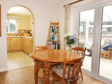 A dining area with a round wooden table and chairs with a vase of flowers by a glass door and a ladder shelf with books at Broadway Lands Farm in Little Dewchurch