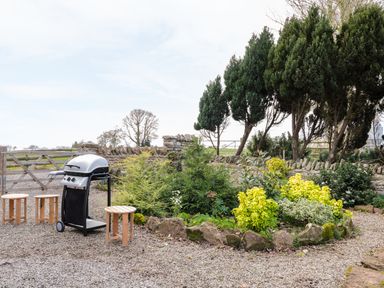 A garden area with a barbecue grill and three wooden stools near green shrubs and trees at Broadway Lands Farm in Little Dewchurch