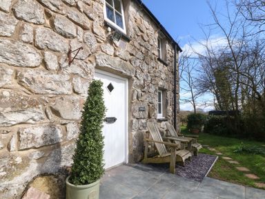 Exterior view of a stone cottage with a white door two wooden chairs and a potted plant at Y Bwthyn Ty Du in Pwllheli