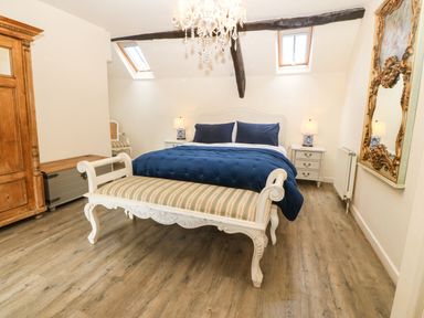 A bedroom with a white bed with blue bedding a striped bench wooden wardrobe two bedside tables with lamps a decorative mirror and wooden flooring at Y Bwthyn Ty Du in Pwllheli