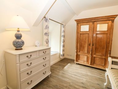 A room with a white chest of drawers with a lamp on it a wooden wardrobe with mirrors on the doors and a window with patterned curtains at Y Bwthyn Ty Du in Pwllheli