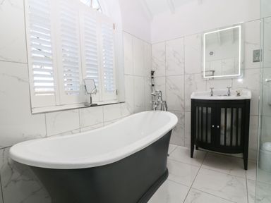 A bathroom with a freestanding bathtub a sink with cabinet a backlit mirror and a window with white shutters at Y Bwthyn Ty Du in Pwllheli