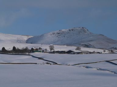 A snow-covered rural landscape with farm buildings and a hill in the background at Woodpecker Lodge in Gargrave