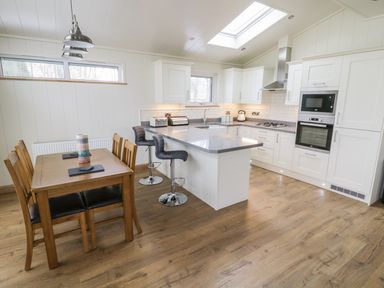 A kitchen with white cabinets and an island with two stools next to a wooden dining table with six chairs at Woodpecker Lodge in Gargrave