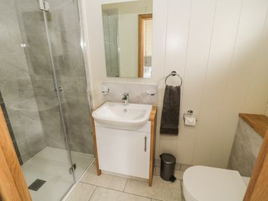 A bathroom with a glass shower, white sink with wooden cabinet, mirror, gray towel, toilet, and small trash can at Woodpecker Lodge in Gargrave