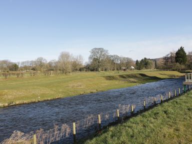 A river flowing beside grassy fields with trees in the background at Woodpecker Lodge in Gargrave