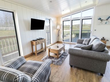 A living room with a grey sofa, plaid armchair, coffee table, TV on the wall, and sliding glass doors at Woodpecker Lodge in Gargrave