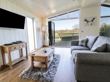 A living room with a grey sofa a wooden coffee table a TV on the wall and a door opening to a deck with a view at Woodpecker Lodge in Gargrave