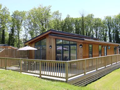 A wooden cabin with a deck and railing surrounded by grass and trees at Woodpecker Lodge in Gargrave