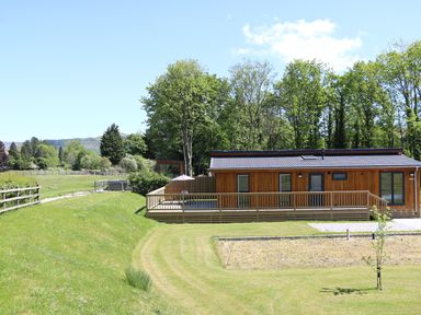 A wooden cabin with a deck and outdoor furniture surrounded by grass and trees at Woodpecker Lodge in Gargrave