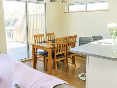 A dining area with a wooden table and four chairs next to a sliding glass door and a white kitchen counter with two gray bar stools at Woodpecker Lodge in Gargrave