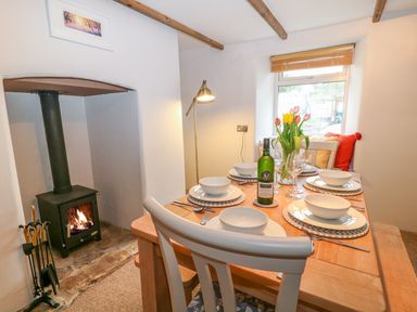 A dining room with a wooden table set for six with plates bowls and glasses a bottle of wine a vase of flowers a wood burning stove and a window with cushions on the windowsill at Blue Anchor in Fraddon