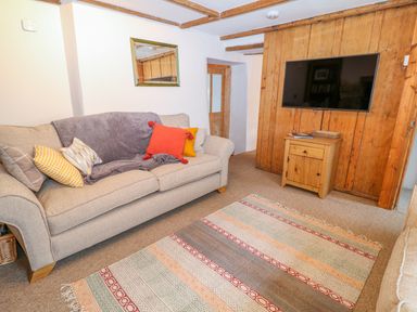 A living room with a sofa with cushions and a throw a patterned rug a wall mounted television and a wooden cabinet at Blue Anchor in Fraddon