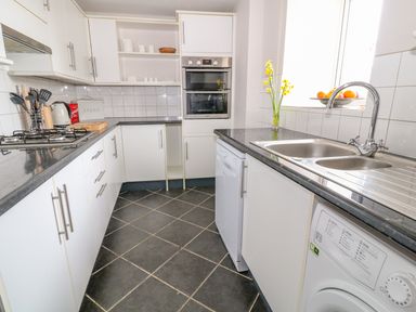 A kitchen with white cabinets, black countertops, a gas stove, double oven, sink, dishwasher, washing machine, and kitchen utensils at Blue Anchor in Fraddon