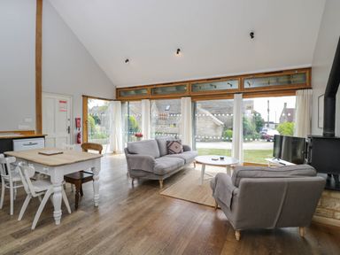 A living room with two gray sofas a white round coffee table and a dining table with chairs near large windows at The Barn at Daubeneys in Colerne