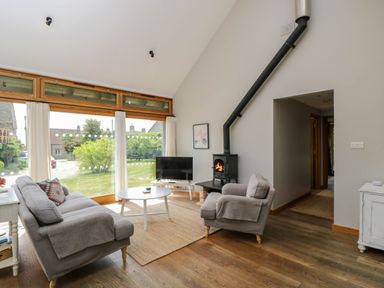 A living room with large windows a sofa armchair coffee table wood stove and television at The Barn at Daubeneys Colerne