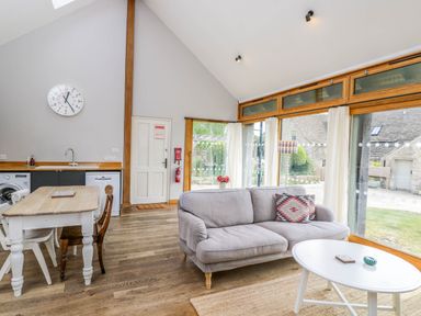 A living area with a gray sofa, white coffee table, dining table with chairs, and large windows at The Barn at Daubeneys in Colerne
