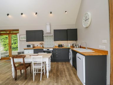 A kitchen with dark cabinets wooden countertops a white table with two chairs a clock on the wall and wooden floor at The Barn at Daubeneys in Colerne