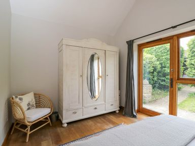 A bedroom with a white wardrobe with mirrored doors a wicker chair wooden floor and glass doors opening to a garden at The Barn at Daubeneys Colerne