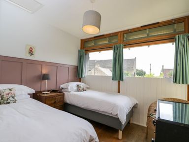 A bedroom with two single beds a wooden nightstand and windows with green curtains at The Barn at Daubeneys in Colerne
