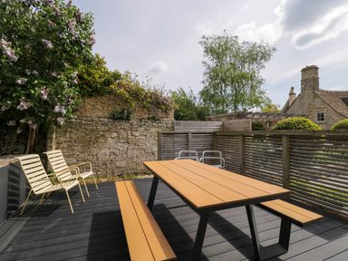 An outdoor patio with wooden picnic table benches and metal chairs surrounded by stone walls and greenery at The Barn at Daubeneys Colerne
