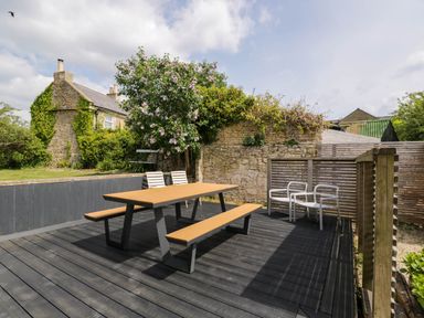 An outdoor patio with a wooden picnic table bench set and metal chairs surrounded by stone walls and plants at The Barn at Daubeneys in Colerne