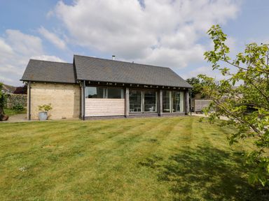 A single story house with stone and wood exterior walls and a lawn with a tree in the foreground at The Barn at Daubeneys in Colerne