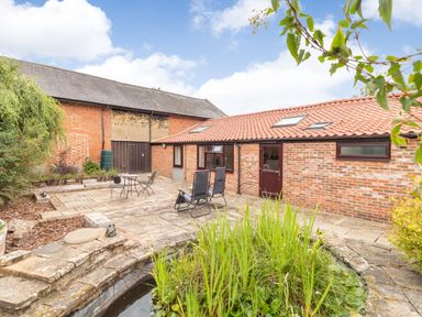 An outdoor patio area with chairs and table next to a pond and brick buildings at The Hen House in Diss near Gissing