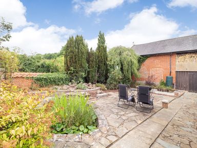 A garden patio with two chairs a small pond tall trees and a brick building at The Hen House in Diss near Gissing