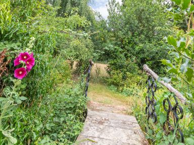 A small stone bridge with metal railings surrounded by green bushes and pink flowers at The Hen House in Diss near Gissing