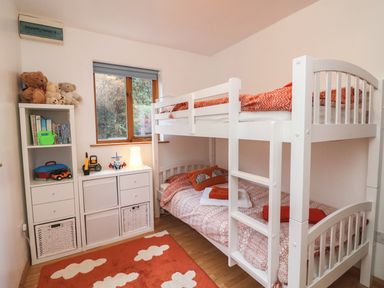 A children's bedroom with white bunk beds a shelving unit with toys and books and a window at Cross Syke Barn in Loxhore near Barnstaple