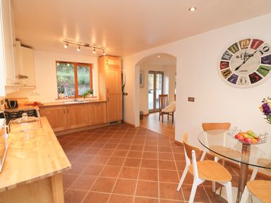 A kitchen with wooden cabinets and a window above the sink a round glass table with wooden chairs and a wall clock at Cross Syke Barn in Loxhore near Barnstaple