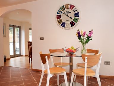 A dining area with a round glass table and four wooden chairs with flowers in a vase and a fruit bowl on the table at Cross Syke Barn in Loxhore near Barnstaple