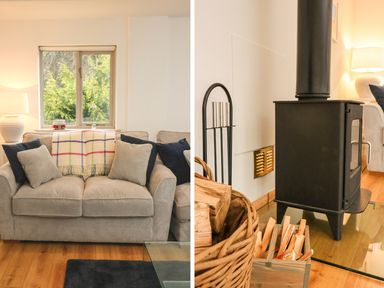 A living room with a beige sofa with cushions and a blanket a window and a lamp next to a black wood stove with firewood and kindling at Cross Syke Barn in Loxhore near Barnstaple
