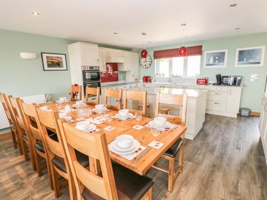 A dining area with a wooden table set for ten and a kitchen with white cabinets and appliances at Harmon Vale in Solva