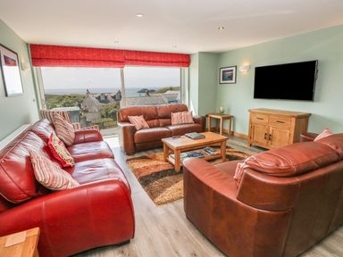A living room with three leather sofas a wooden coffee table a flat screen TV on the wall large windows with red blinds and a view of houses and the sea at Harmon Vale in Solva