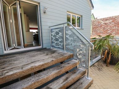An outdoor wooden staircase with metal fish design railing leading to an open doorway of a light blue house with potted plants on a paved area at Harmon Vale in Solva