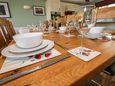 A dining table set with white bowls, plates, wine glasses, cutlery and poppy flower placemats in a kitchen at Harmon Vale in Solva