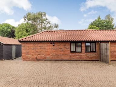 A red brick building with a tiled roof and three windows facing a paved open area with trees in the background at The Nest Box in Diss