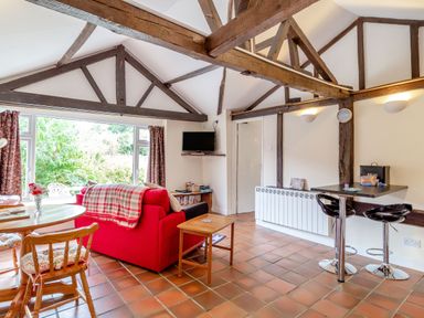 A living area with a red sofa a wooden round table wooden chairs a small wooden side table a wall-mounted TV and a black breakfast bar with two stools at The Nest Box in Diss