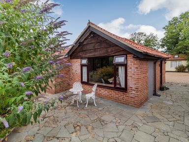 A small brick building with a tiled roof and large windows next to a stone paved patio with white metal table and chairs and a flowering bush at The Nest Box in Diss
