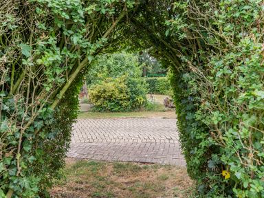A garden archway made of green leafy plants with a brick pathway and bushes beyond at The Nest Box in Diss