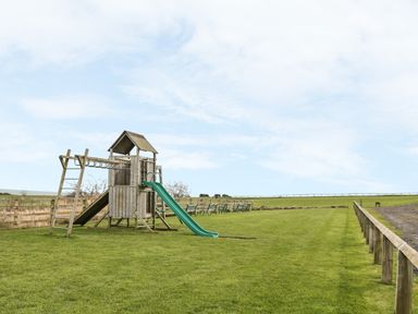 A wooden playground structure with slides and monkey bars on a grass field with benches in the background at St Dolmen in Embleton