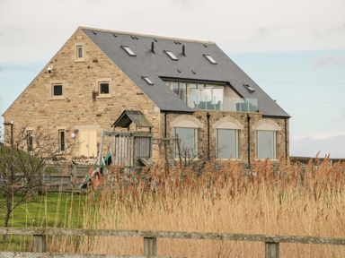 A stone house with multiple windows and a glass balcony with a playground and tall grass in front at St Dolmen in Embleton
