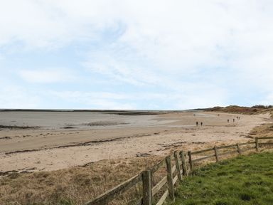 A sandy beach with a wooden fence and people walking along the shore at St Dolmen in Embleton