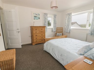 A bedroom with a bed, wooden chest of drawers, chair with a cushion, wicker basket, and windows with curtains at 54 The Moorings in St Dogmaels