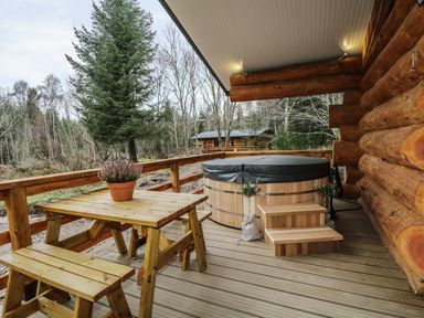 An outdoor wooden deck with a picnic table and benches a covered hot tub and a log cabin wall at Moorhen Lodge in North Kessock