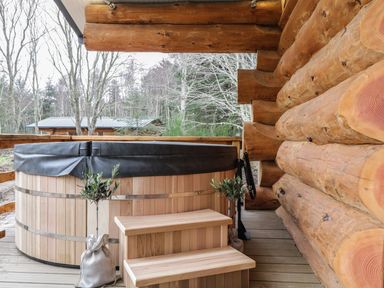 An outdoor wooden hot tub with steps next to a log cabin wall at Moorhen Lodge in North Kessock