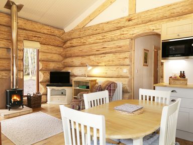 A dining area with a wooden table and white chairs next to a living space with a plaid armchair and a wood stove in a log cabin at Moorhen Lodge in North Kessock